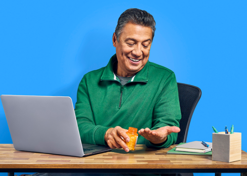 Man sits at desk holding medication in left hand blue background